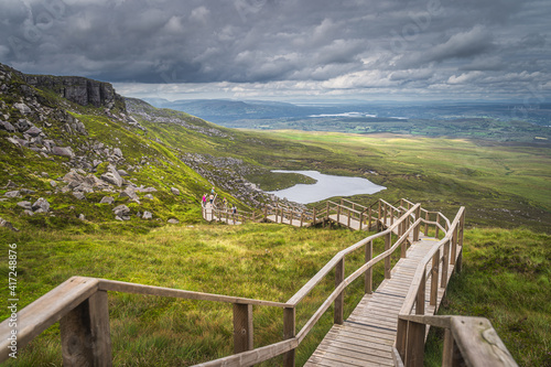 People enjoying a walk on steep stairs of wooden boardwalk in Cuilcagh Mountain Park with a view on lake and valley below, Northern Ireland photo