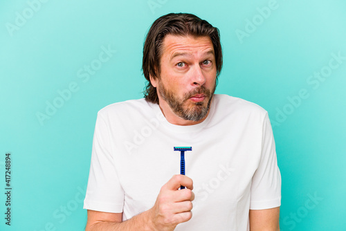 Middle age dutch man holding a razor blade isolated on blue background shrugs shoulders and open eyes confused.