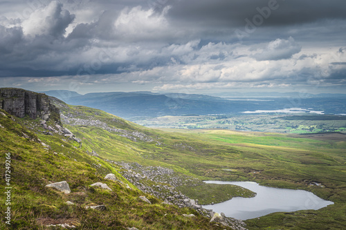 Green fields and large boulders illuminated by sunlight in Cuilcagh Mountain Park with small lakes in valley below, Northern Ireland