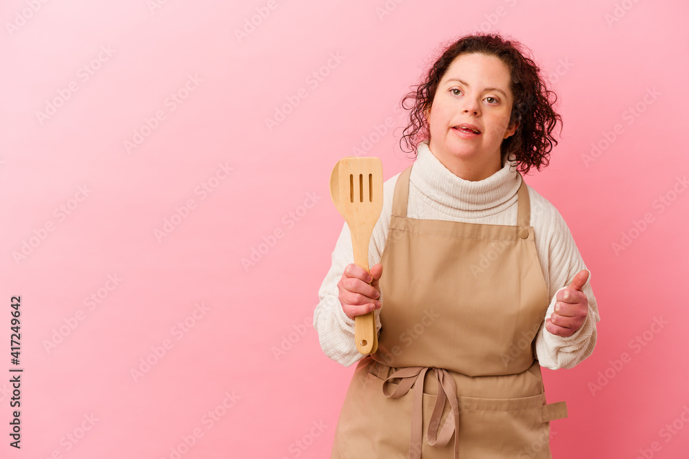 Woman with Down syndrome cooking at home isolated on pink background smiling and raising thumb up Woman with Down syndrome cooking at home isolated on pink background smiling and raising thumb up