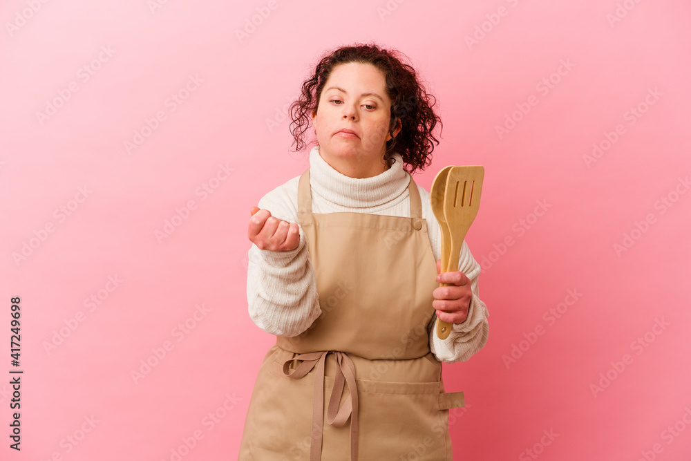 Woman with Down syndrome cooking at home isolated on pink background showing fist to camera, aggressive facial expression. Woman with Down syndrome cooking at home isolated on pink background showing fist to camera, aggressive facial expression.
