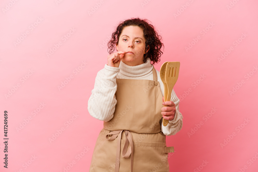 Woman with Down syndrome cooking at home isolated on pink background with fingers on lips keeping a secret. Woman with Down syndrome cooking at home isolated on pink background with fingers on lips keeping a secret.