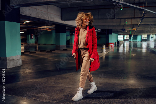 Young smiling fashionable woman with curly hair walking in underground garage and looking away.