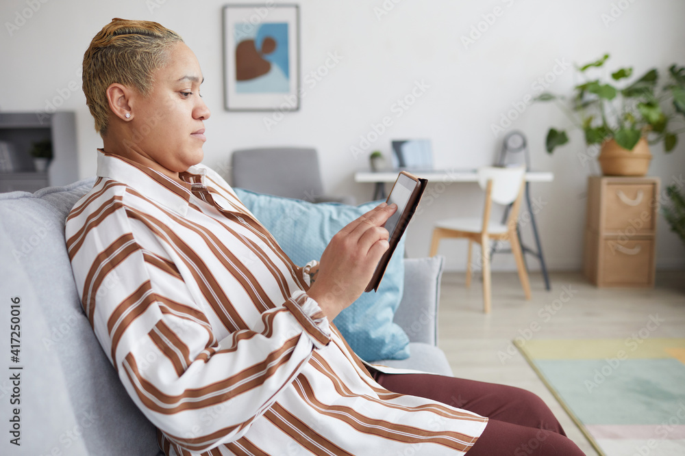 Side view portrait of modern mixed-race woman holding digital tablet and using online services while sitting on sofa at home, copy space