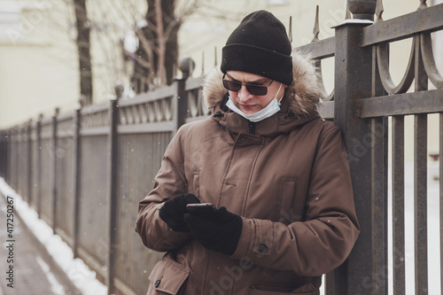 Portrait of young man in casual winter clothes with glasses and medical mask on walk city. An attractive teenager stands in old town looking mobile phone and emotion unexpected email on frosty day