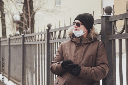 Portrait of young man in casual winter clothes with glasses and medical mask on walk city. An attractive teenager stands in old town looking mobile phone and emotion unexpected email on frosty day © Alex Vog