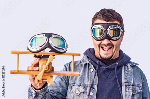 Fashionable fooling young man in pilot glasses with a wooden airplane in his hand against the blue sky outdoors. Concept purpose, adventure, travel, freedom. Mens fashion. Spring clothes. Jeans, denim