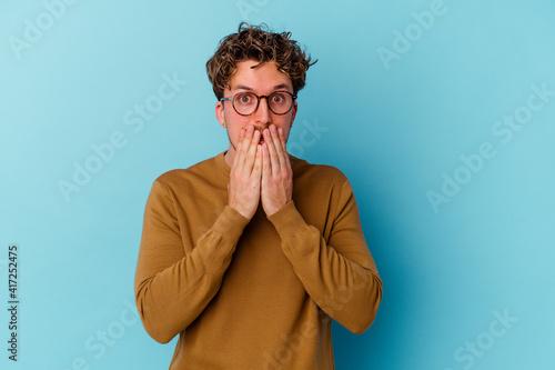 Young caucasian man wearing eyeglasses isolated on blue background shocked  covering mouth with hands  anxious to discover something new.