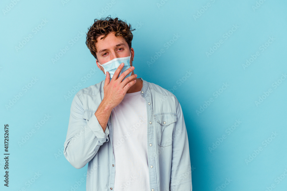 Young caucasian man wearing an antiviral mask isolated on blue background yawning showing a tired gesture covering mouth with hand. Young caucasian man wearing an antiviral mask isolated on blue background yawning showing a tired gesture covering mouth with hand.