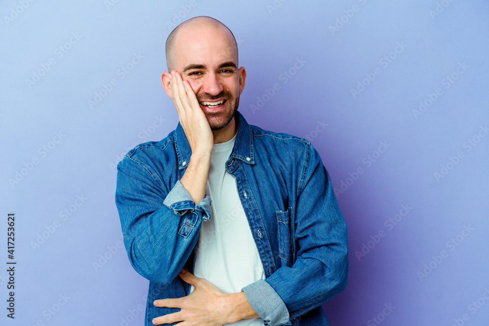 Young caucasian bald man isolated on purple background laughs happily and has fun keeping hands on stomach. Young caucasian bald man isolated on purple background laughs happily and has fun keeping hands on stomach.
