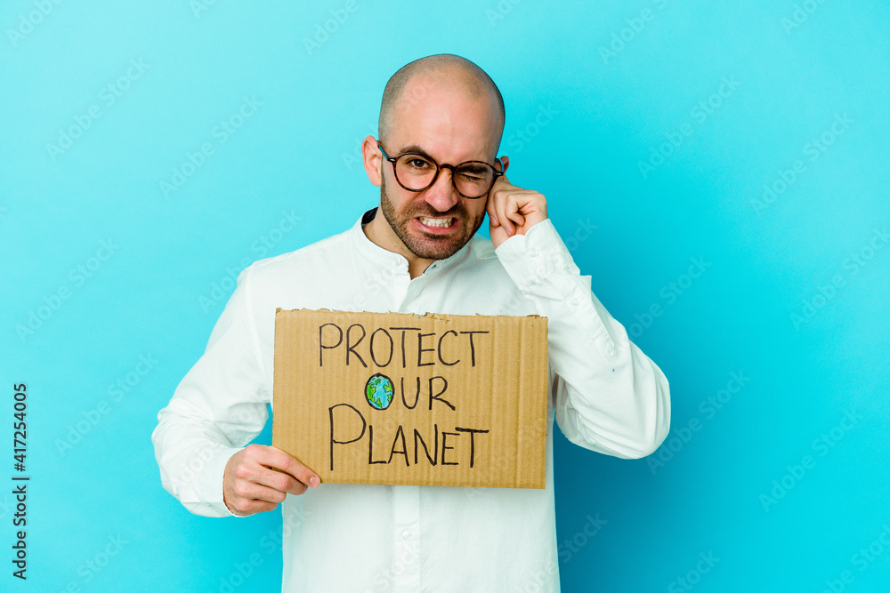 Young caucasian bald man holding a protect our planet placard isolated on purple background covering ears with hands. Young caucasian bald man holding a protect our planet placard isolated on purple background covering ears with hands.