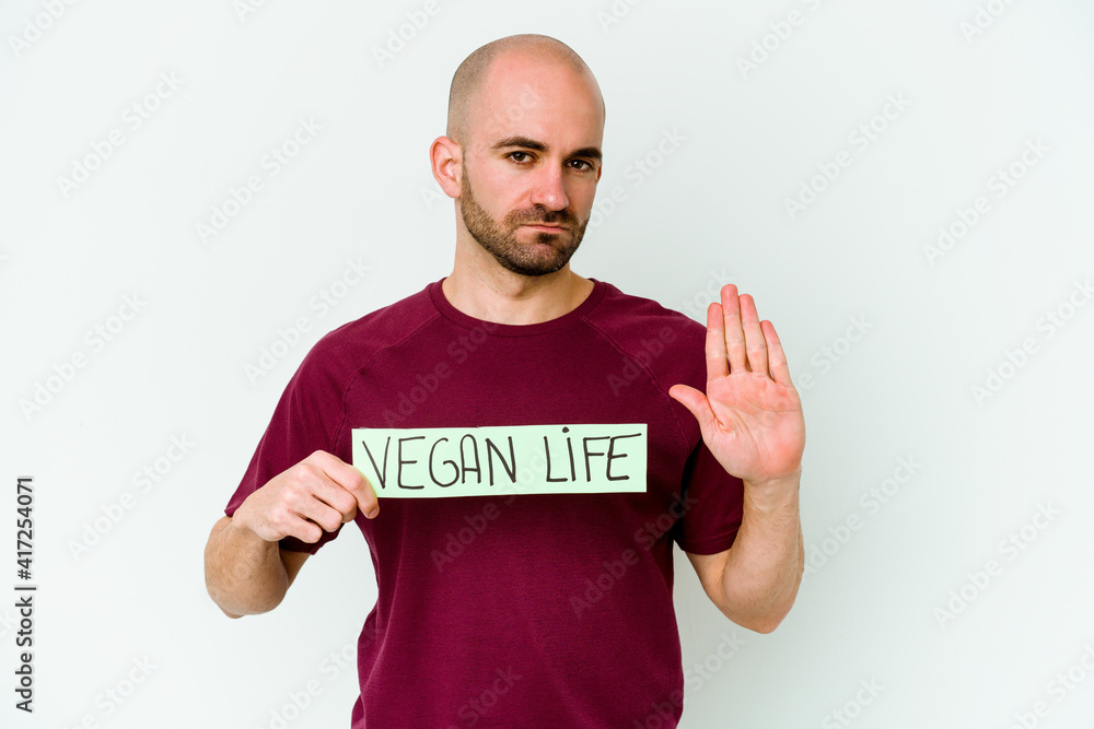 Young caucasian bald man holding a Vegan life placard isolated on purple background standing with outstretched hand showing stop sign, preventing you. Young caucasian bald man holding a Vegan life placard isolated on purple background standing with outstretched hand showing stop sign, preventing you.
