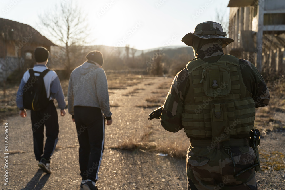 Group of two migrants held by soldier at border patrol on the road in nature in sunny day - Back view of two unknown men facing armed man in uniform on road after being arrested for illegal activity Group of two migrants held by soldier at border patrol on the road in nature in sunny day - Back view of two unknown men facing armed man in uniform on road after being arrested for illegal activity