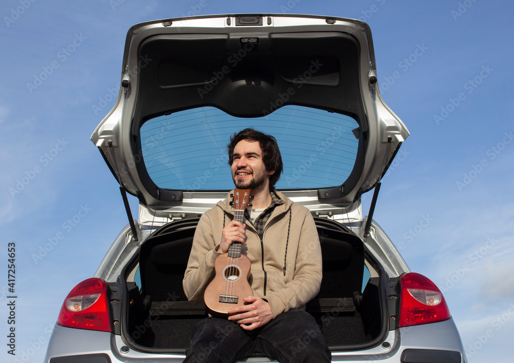 Naklejka premium Smiled young man with ukulele in his hands sitting in the trunk. Young man playing ukulele