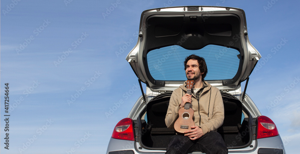 Smiled young man with ukulele in his hands sitting in the trunk. Young man playing ukulele. Copy space