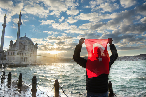 Man hold Turkish flag. Sunrise near Bosphorus bridge (aka: 15 July Martyrs Bridge Turkish: 15 Temmuz Sehitler Koprusu) and Imperial Mosque of Sultan Abdulmecid in Ortakoy, Istanbul, Turkey. Patriotism photo