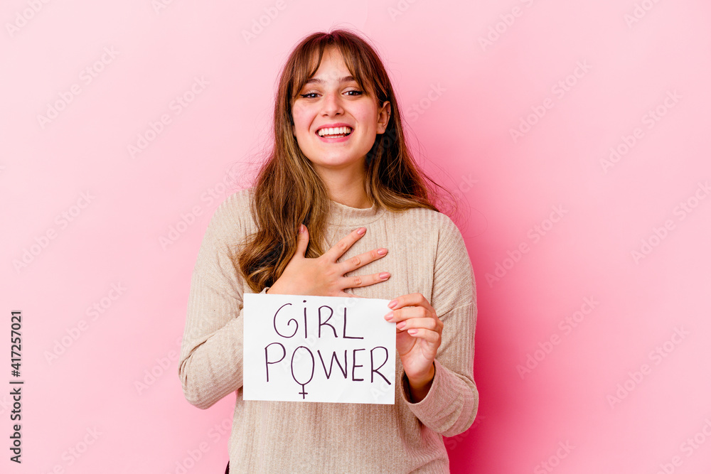 Young caucasian woman holding a girl power placard isolated on pink background laughs out loudly keeping hand on chest. Young caucasian woman holding a girl power placard isolated on pink background laughs out loudly keeping hand on chest.