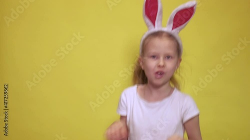 Cute little girl with bunny ears looking at camera and jumping while pretending to be rabbit during Easter celebration against yellow background