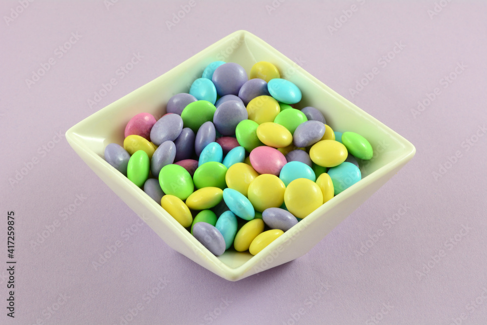 Pastel Easter candy coated milk chocolate candy pieces in white candy dish on lavender background Pastel Easter candy coated milk chocolate candy pieces in white candy dish on lavender background