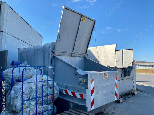 Grauer Container, ein Flügel weit geöffnet für plalttgepresste Pfandplastikflaschen in Plastikbeutel verpackt. Betonwand und strahlend blauer Himmel drum herum. photo