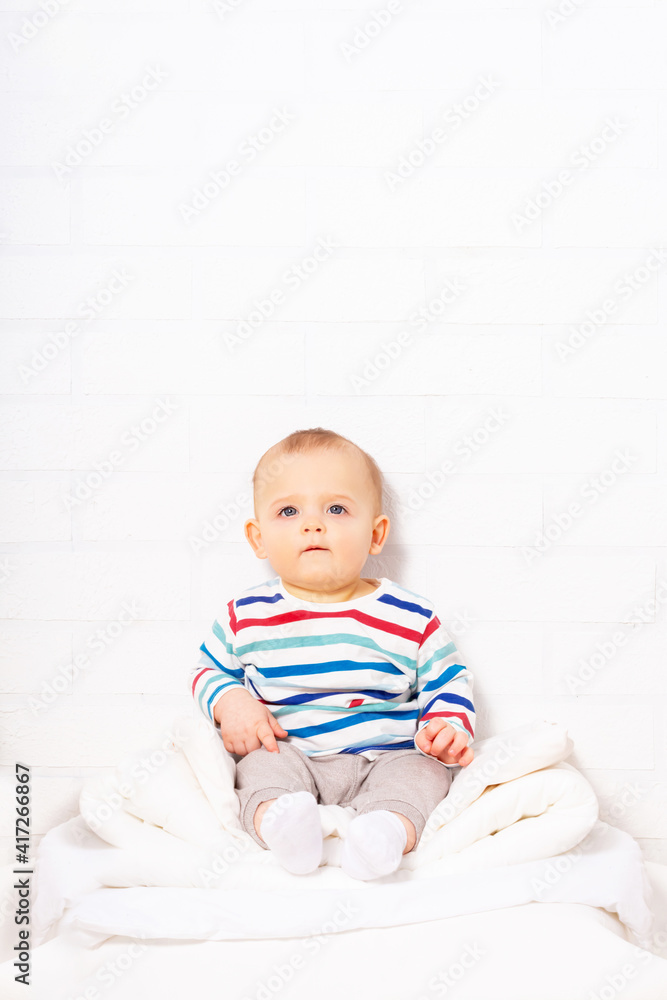 Funny caucasian baby boy in striped t-shirt sitting on pillow mountain. White brick wall background with copy space. Funny caucasian baby boy in striped t-shirt sitting on pillow mountain. White brick wall background with copy space.