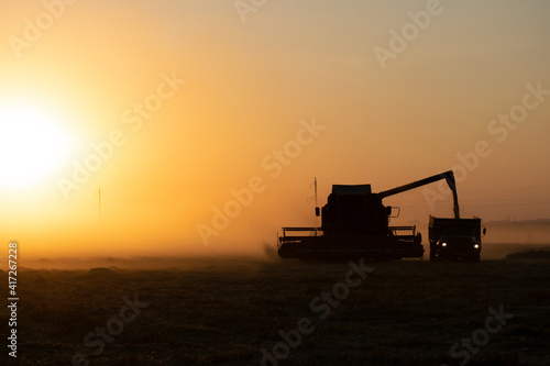 The harvester is pouring grain into the flatbed truck. There is a lot of dust around in the evening.