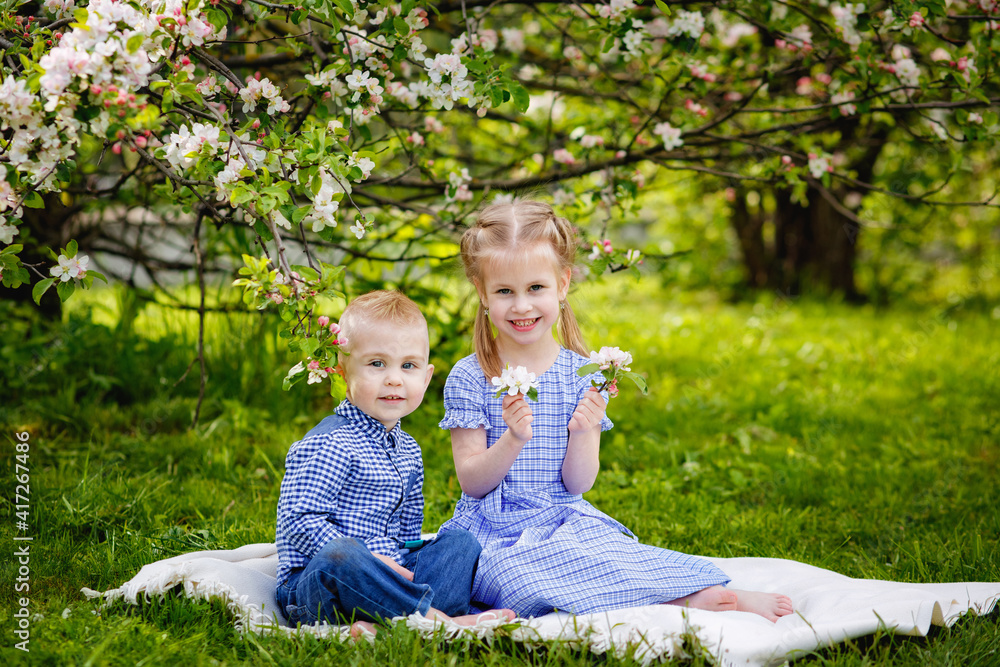 Cute kids girl and boy sit on the grass under an apple tree in a spring blooming garden. Brother and sister on a picnic in the spring. Cute kids girl and boy sit on the grass under an apple tree in a spring blooming garden. Brother and sister on a picnic in the spring.