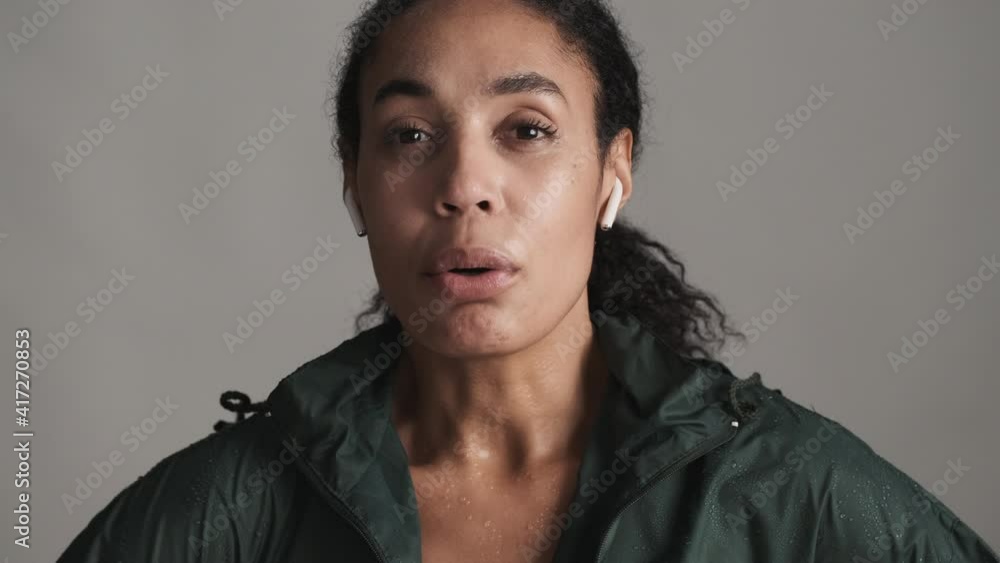 Young tired African American woman in wireless earphones hard breathing after run but smiling on camera looking happy over white background. Sport lover Young tired African American woman in wireless earphones hard breathing after run but smiling on camera looking happy over white background. Sport lover