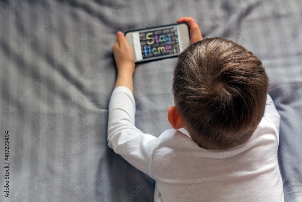 Kid boy looking information text at Internet on mobile phones (smartphone) lying in bed during quarantine. Digital technology, gadget addiction Kid boy looking information text at Internet on mobile phones (smartphone) lying in bed during quarantine. Digital technology, gadget addiction