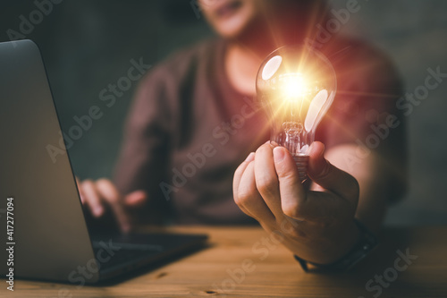 Quadro su tela Woman hand holding light bulb and using laptop on wooden table