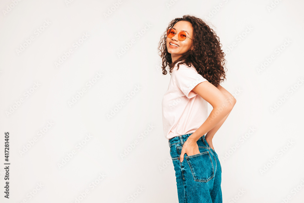 Beautiful black woman with afro curls hairstyle.Smiling model in white trendy jeans clothes. Sexy carefree female posing on grey background in studio in sunglasses. Tanned and cheerful