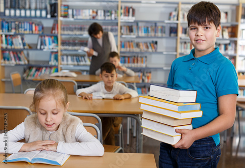 Smiling intelligent boy holding pile of books while standing in modern school library