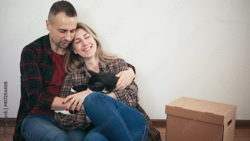 An adult couple hugging a cat while sitting on the floor by the white wall in the living room next to the cardboard box after moving into their new apartment. An adult couple hugging a cat while sitting on the floor by the white wall in the living room next to the cardboard box after moving into their new apartment.