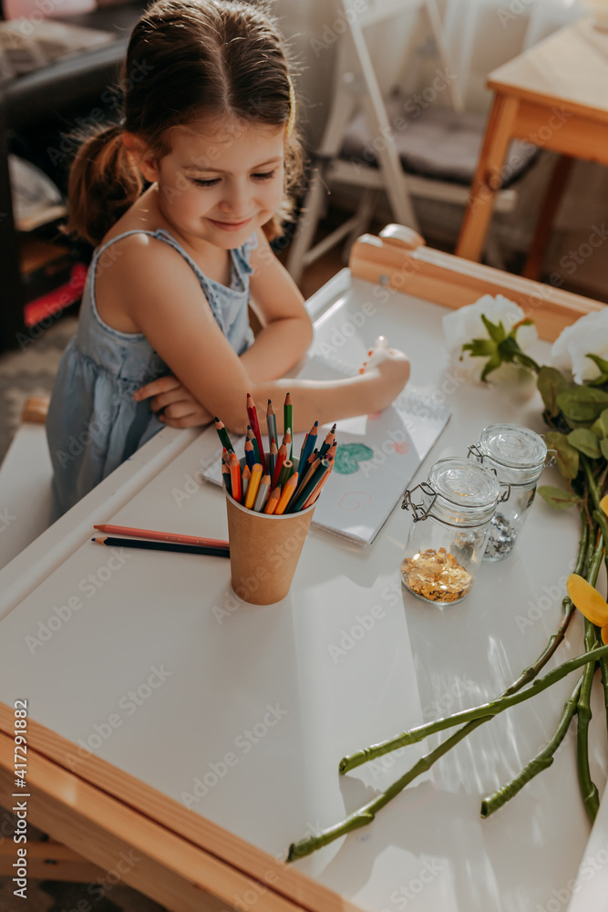 Little girl drawing hearts on white paper. Happy mother's day card with girl sitting near white table. Kid makes homework with pencils. Girl with flowers.