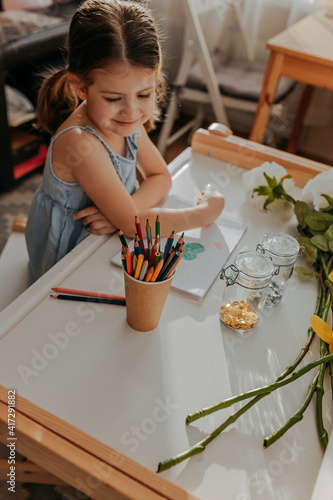 Obraz na płótnie Little girl drawing hearts on white paper. Happy mother's day card with girl sitting near white table. Kid makes homework with pencils. Girl with flowers. w salonie