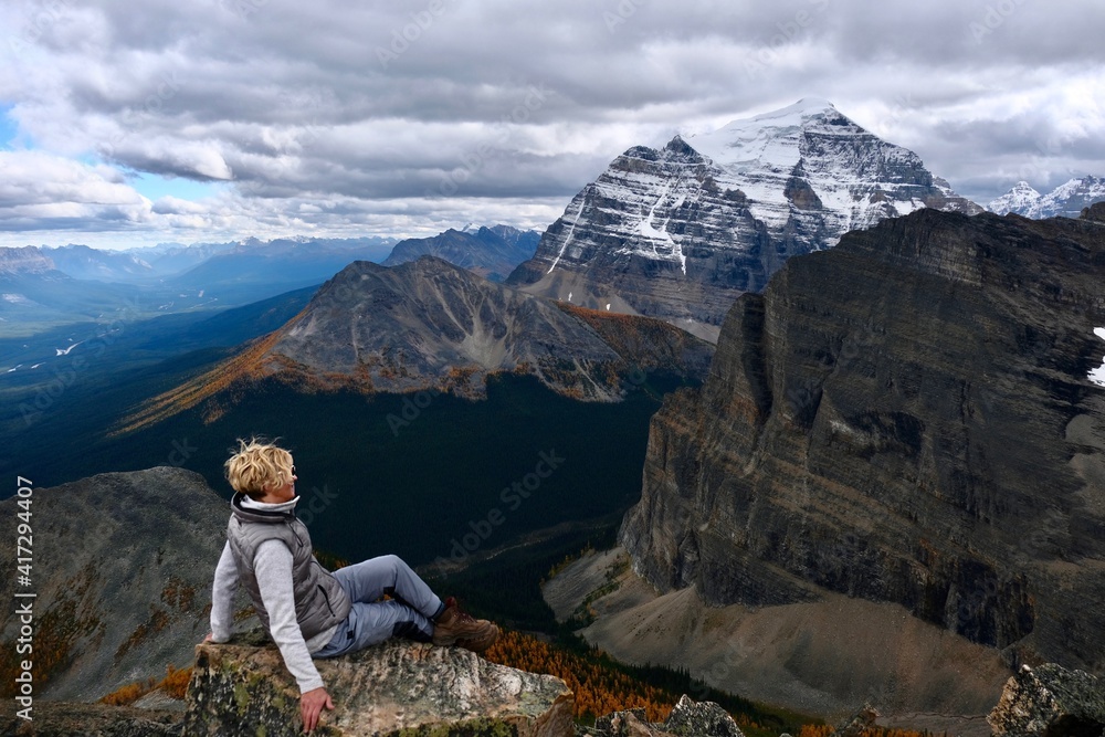 Woman hiker sitting on mountain top enjoing view of mountain valley with yellow larch trees from above. Lake Louise area. Banff National park. Alberta. Canada.