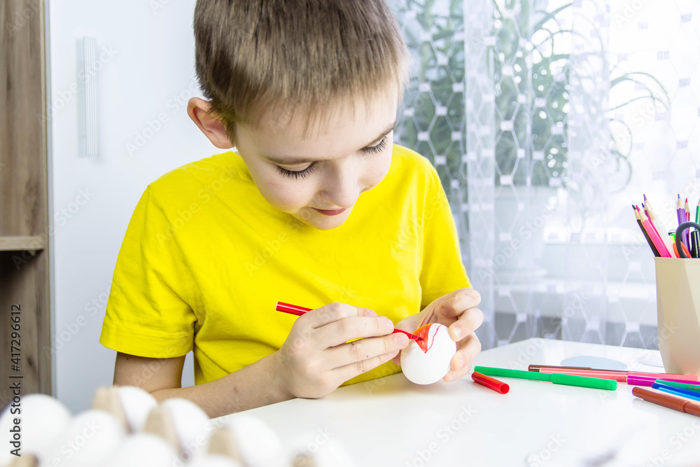 A boy in a yellow T-shirt sits at a table and paints a white egg with colored markers. The concept of preparation for Easter. A boy in a yellow T-shirt sits at a table and paints a white egg with colored markers. The concept of preparation for Easter.