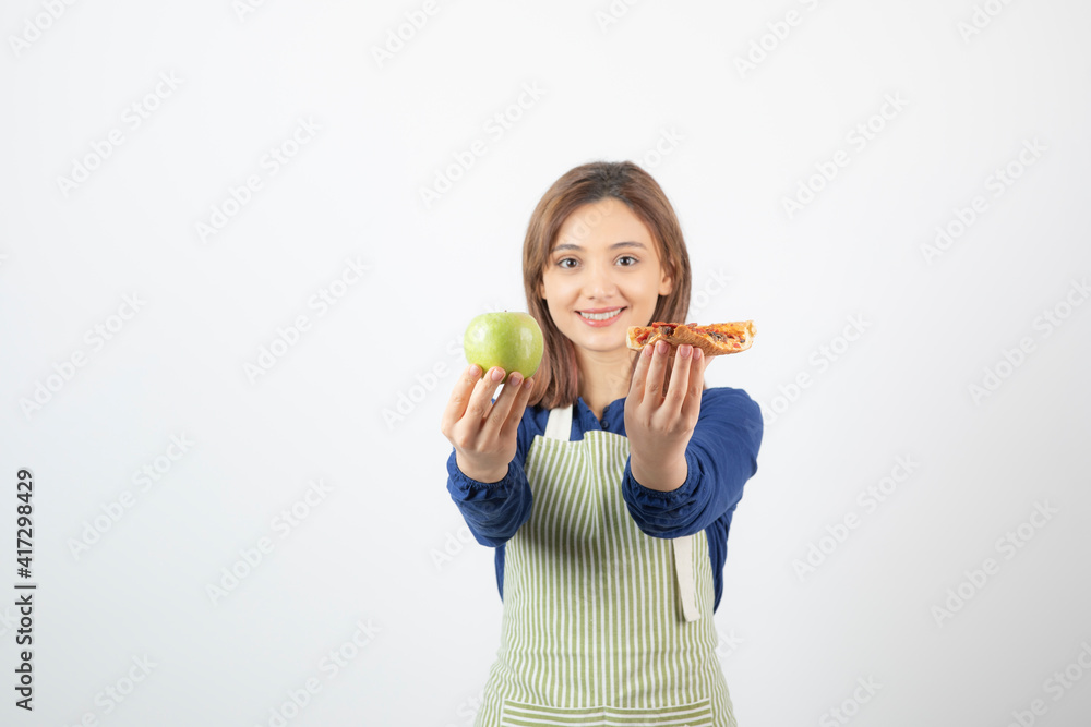 Image of woman in apron trying to choose what to eat apple or pizza Image of woman in apron trying to choose what to eat apple or pizza