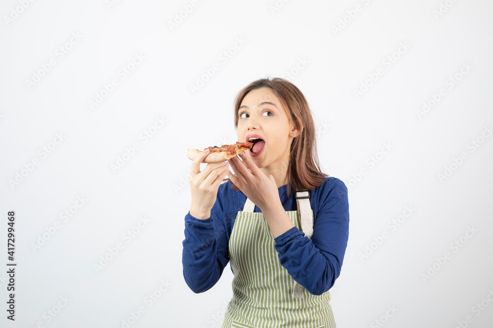Portrait of young woman in apron eating pizza on white background