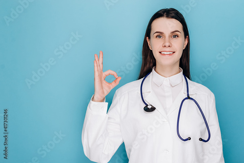Happy female doctor show okay gesture  smiling at camera with optimistic satisfied expression  wears white coat and stethoscope  isolated on blue background with copy space. Healthcare workers concept