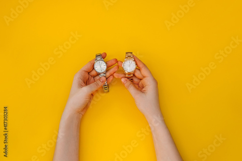Female hands holding two wrist watches, golden and silver, on yellow background.