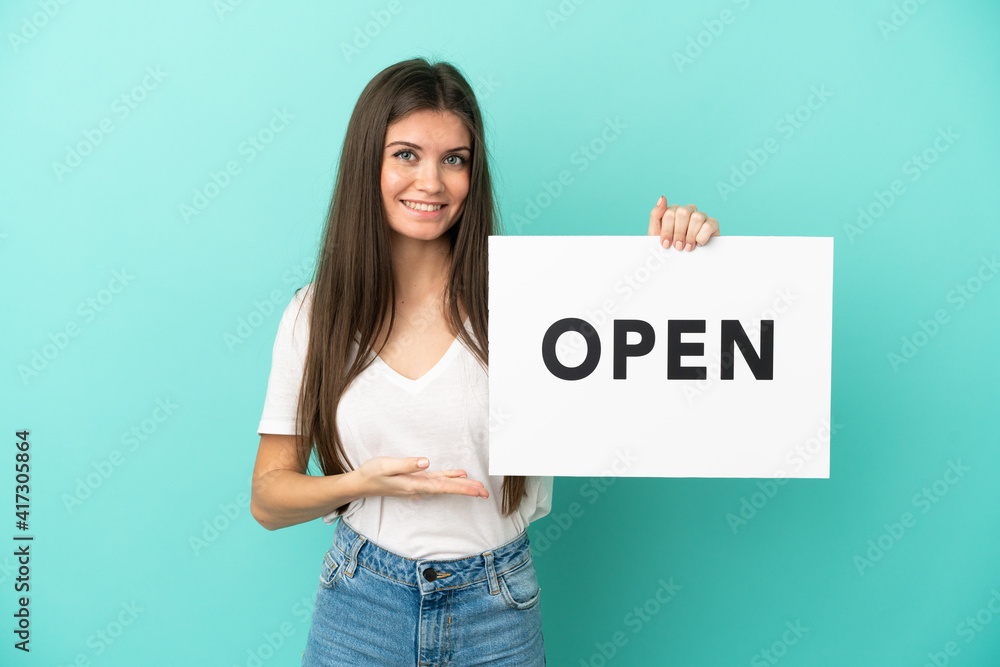 Young caucasian woman isolated on blue background holding a placard with text OPEN and pointing it Young caucasian woman isolated on blue background holding a placard with text OPEN and pointing it