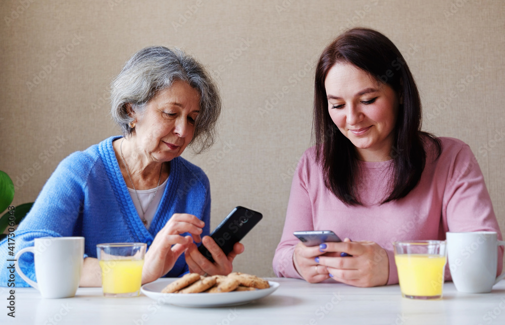 An elderly mother and an adult daughter use phones to view content on social networks - Grandmother and granddaughter are sitting in a cozy house and watching photos or videos on the phone