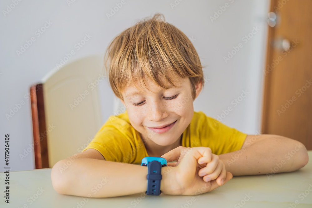 Little boy sitting at the table and looking smart watch. Smart watch for baby safety. The child makes school lessons, listening to music, calling friends Little boy sitting at the table and looking smart watch. Smart watch for baby safety. The child makes school lessons, listening to music, calling friends