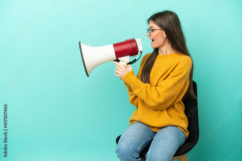 Young caucasian woman sitting on a chair isolated on blue background shouting through a megaphone Young caucasian woman sitting on a chair isolated on blue background shouting through a megaphone