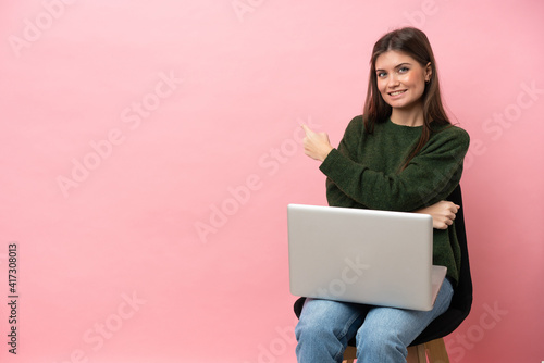 Young caucasian woman sitting on a chair with her laptop isolated on pink background pointing back