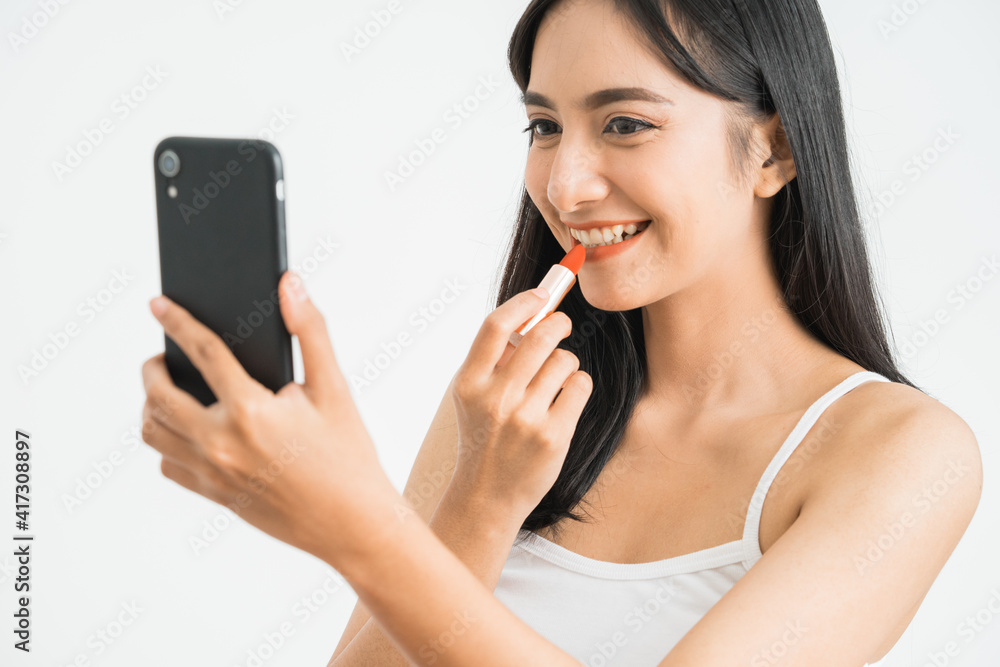 beautiful woman applying lipstick while holding her mobile phone as a mirror over white background. asian female blogger beautiful woman applying lipstick while holding her mobile phone as a mirror over white background. asian female blogger