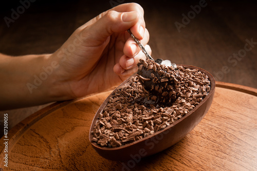 Hand sticking a metallic spoon in a stuffed chocolate easter egg with grated chocolate on the top on a wooden stand on a wooden table. 
