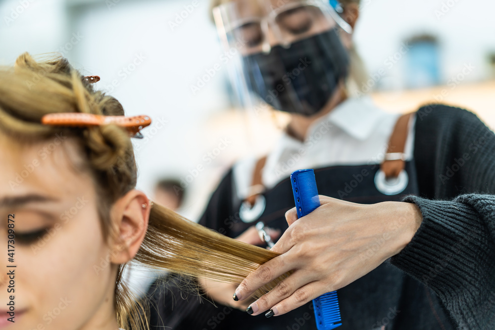 Fototapeta premium Asian professional female stylist cutting Caucasian woman's hair in salon. The man wearing mask and face shield to prevent from coronavirus infection during pandemic. Beauty salon business concept.