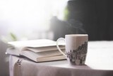 Side view of open book and cup lying on table with white tablecloth. Morning mood, studying, reading, relaxing